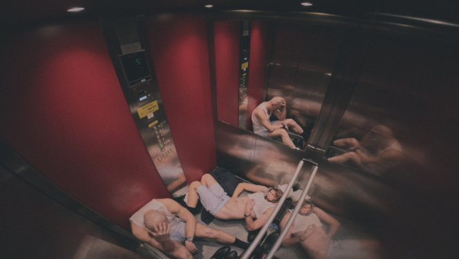 Stopkadrs no filmas “No No Place Hotel“, divi cilvēki liftā sēž uz grīdas
