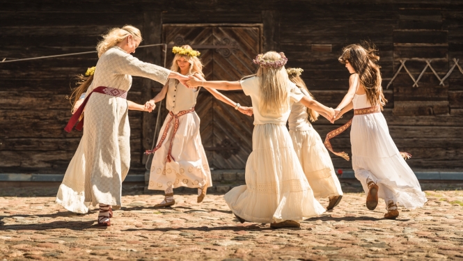 Piecās dejotājas tautiskos tērpos griež danci pagalmā