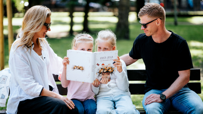 Ģimene - tētis, mamma un divas meitas - brīvā dabā parkā uz soliņa saulainā laikā lasa grāmatu "Atrastā Latvija".