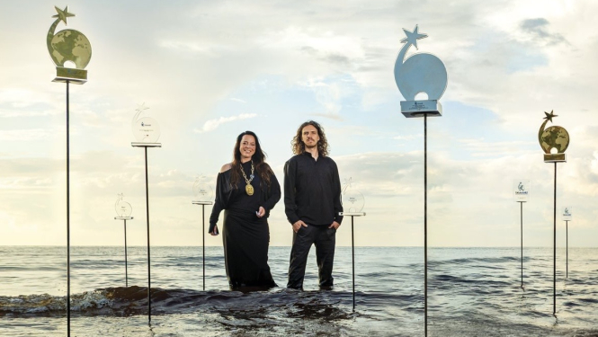 Radošās apvienības “Skudras Metropole” vadītāja un radošā direktore Gundega Skudriņa un dizainers Mārcis Ziemiņš stāv jūrā līdz ceļiem, kur apkārt viņiem uz statīviem novietotas gadu gaitā iegūtās balvas