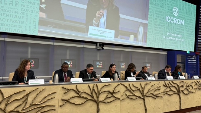 Astoņi cilvēki pie sanāksmju galda, fonā panelis, kur tuvplānā redzams runātājs