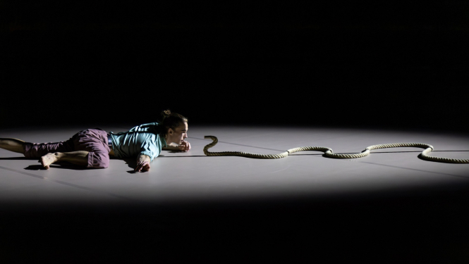 Dejas izrāde, dejotāja pozā uz grīdas, priekšā izlocīta virve