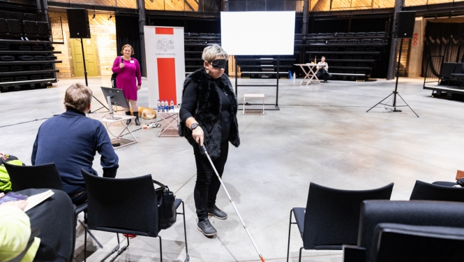 Cilvēks ar spieķi auditorijas priekšā demonstrē piekļūstamības izaicinājumus