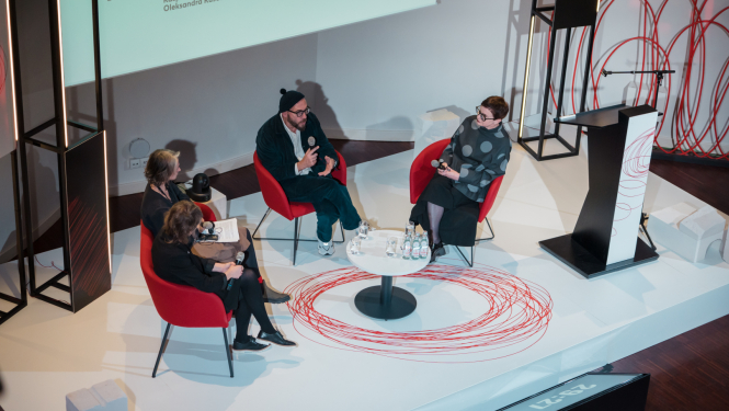 Četri cilvēki sēž modernā konferenču telpā apaļajā diskusijā
