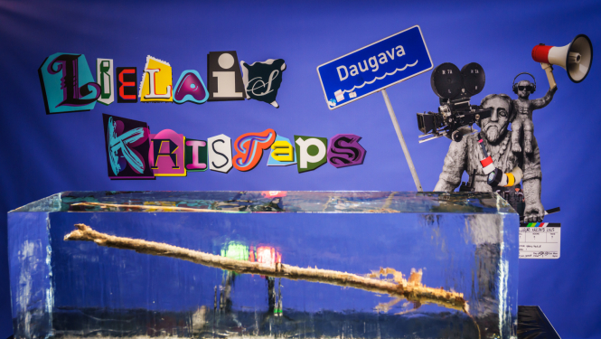 Nacionālās kino balvas „Lielais Kristaps 2025” vizuālā identitāte - baneris ar Lielā Kristapa statueti un ledus gabalā iesalusi koka nūja