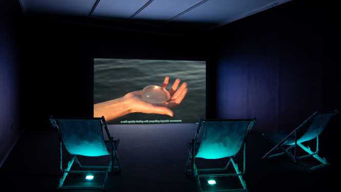 Tumša izstāžu zāle, trīs krēsli, priekšplānā ekrāns ar foto, kur redzama plauksta ar gaišu objektu tajā