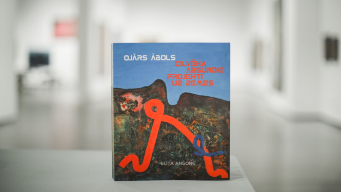 Grāmata “Ojārs Ābols. Cilvēka absurdie projekti uz Zemes” uz gleznotājam veltītās izstādes fona Latvijas Nacionālajā mākslas muzejā