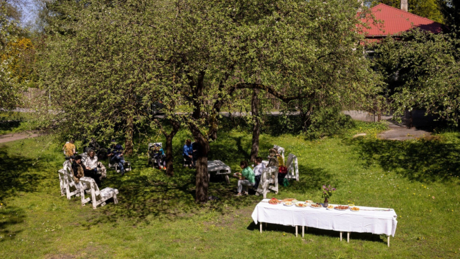 Cilvēki pulcējušies pie balti klāta galda Ojāra Vācieša muzeja dārzā