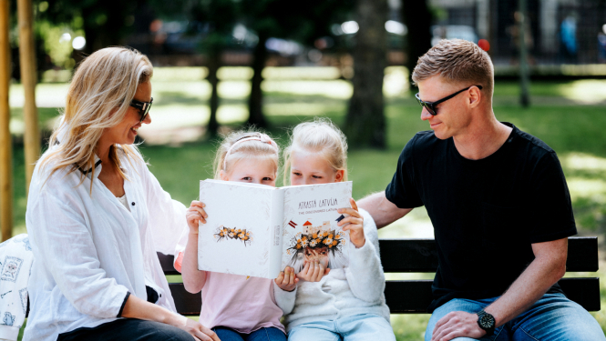 Ģimene - tētis, mamma un divas meitas - brīvā dabā parkā uz soliņa saulainā laikā lasa grāmatu "Atrastā Latvija".