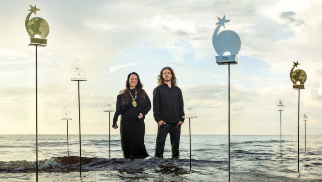 Radošās apvienības “Skudras Metropole” vadītāja un radošā direktore Gundega Skudriņa un dizainers Mārcis Ziemiņš stāv jūrā līdz ceļiem, kur apkārt viņiem uz statīviem novietotas gadu gaitā iegūtās balvas