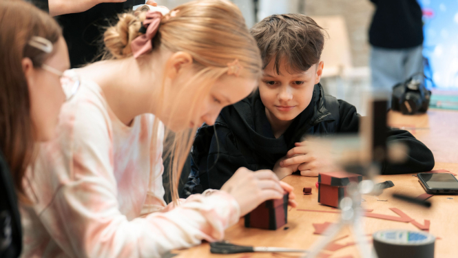 Bērni sēž pie galda, veido kastītes biedrības “ISSP” darbnīcā “Radošā fotogrāfijas laboratorija”