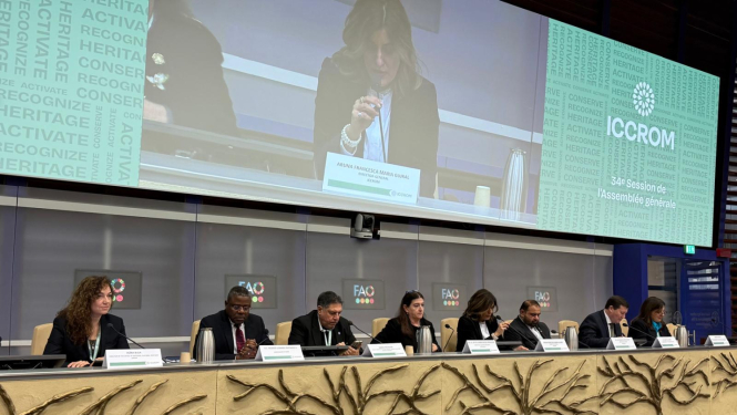 Astoņi cilvēki pie sanāksmju galda, fonā panelis, kur tuvplānā redzams runātājs