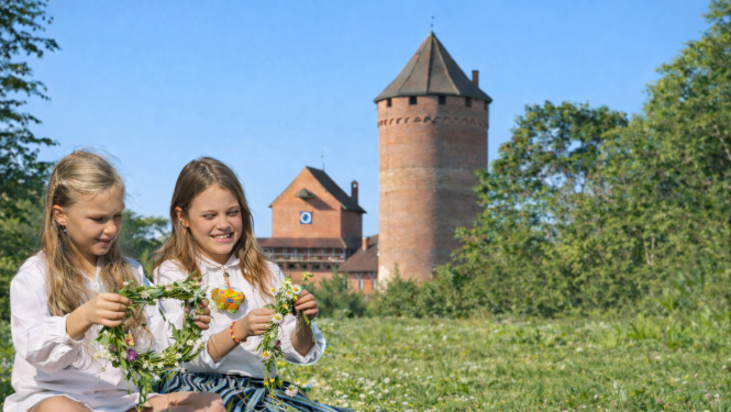 Divas meitenes pin ziedu vainagus pļavā, fonā Turaidas pils