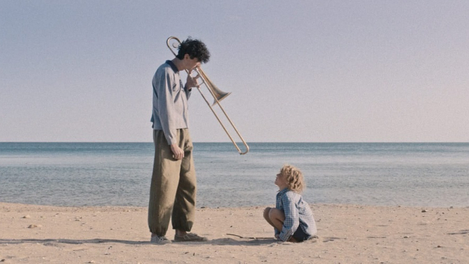 Kadrs no filmas: jūras krastā divi cilvēki - piaugušais ar trombonu rokās un bērns, kas spēlējas smiltīs