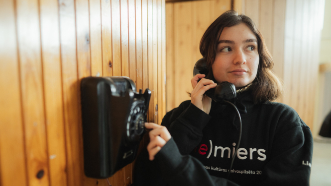 Ilustratīvs attēls ar meiteni, kura stāv pie senlaicīga telefona ar telefona klausuli pie auss un griež ciparnīcu