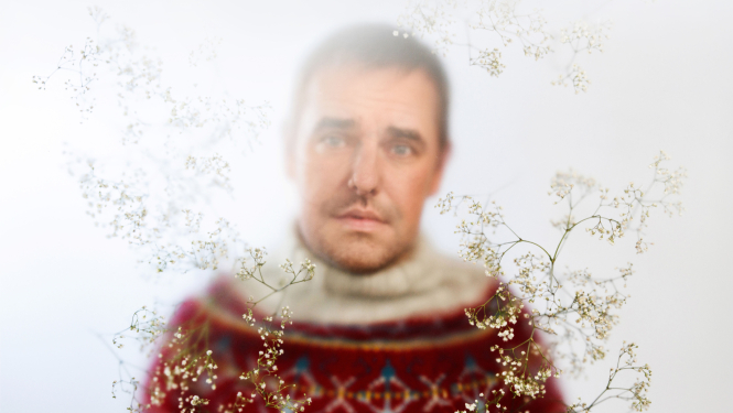 Izrādes “Egles stāsti” publicitātes attēls - neass aktiera portrets ar izžāvētiem pļavas augu elementiem attēla priekšplānā