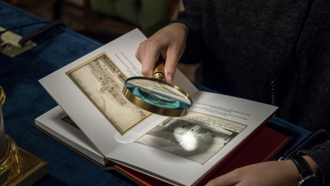 Cilvēka roka tur palielināmo stiklu, kas vērsts pret tekstu grāmatā