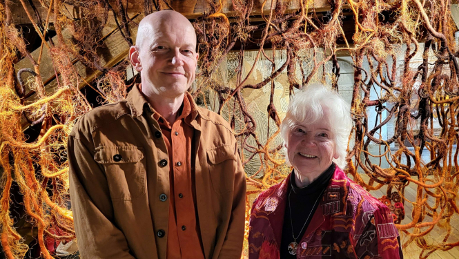Tekstilmāksliniece Vija Jansone un mākslinieks Kaspars Lielgalvis izstādē “Uzkāpt kalnā”, fonā tekstilmākslas darbs