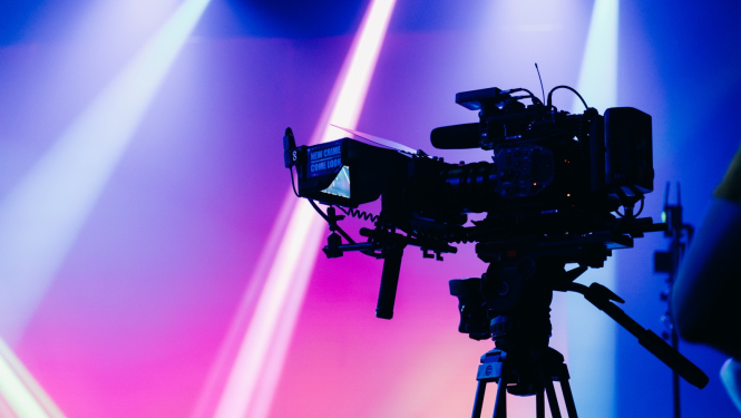 Latvijas Sabiedriskā medija publicitātes attēls. Filmēšanas kamera uz koši apgaismota fona.