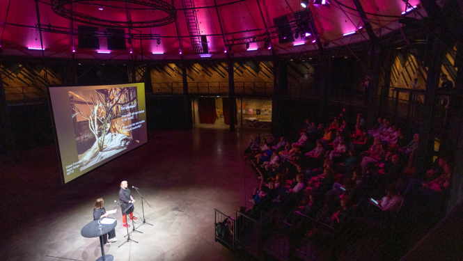 Konferences norise Rīgas cirka arēnā. Divi dalībnieki uzstājas, fonā ekrāns, tribīnēs sēž publika