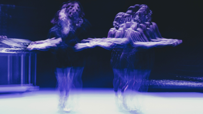 Divi izplūduši dejotāju tēli uz skatuves zilgani violetā gaismā