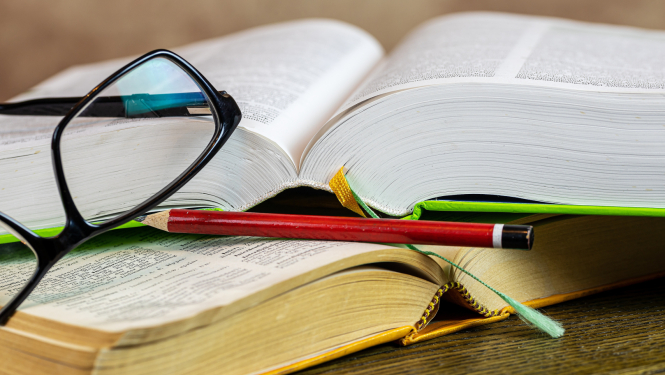 Uz divām atvērtām grāmatām novietots zīmulis un brilles.