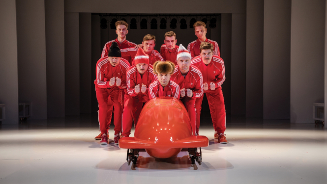 Liepājas teātra izrāde “Latviešu raķetes”, uz skatuves bobsleja kamanas un grupa ar sarkanas krāsas sporta formās tērptiem aktieriem