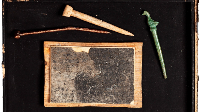 Izstādes “Viduslaiku Rīgas rakstāmlietas” publicitātes attēls. Kompozīcijā vaska tāfelīte un rakstāmirbuļi – stili.