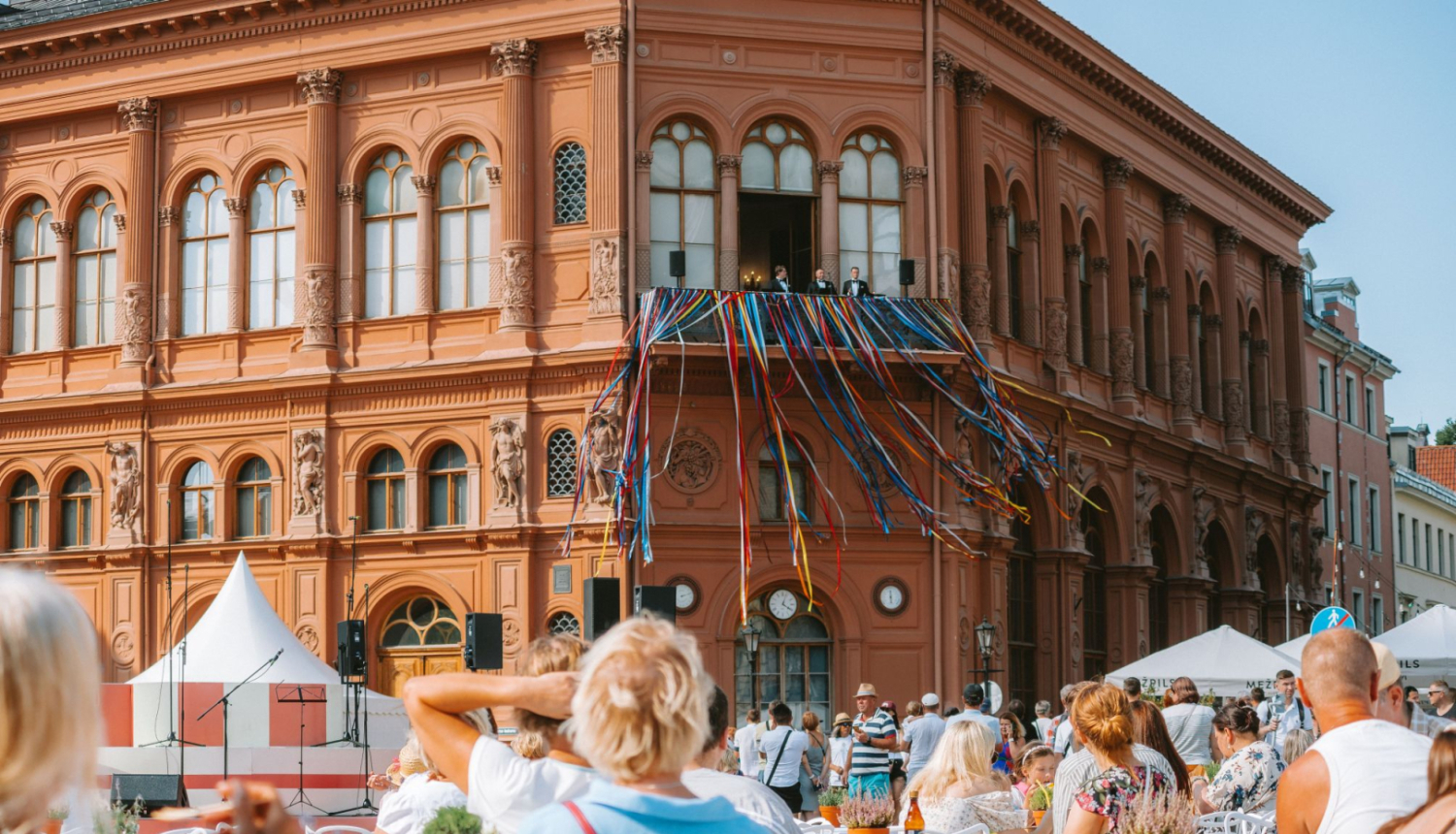 Mākslas muzeja Rīgas Birža fasāde skatā no Doma laukuma, priekšplānā cilvēki sēž