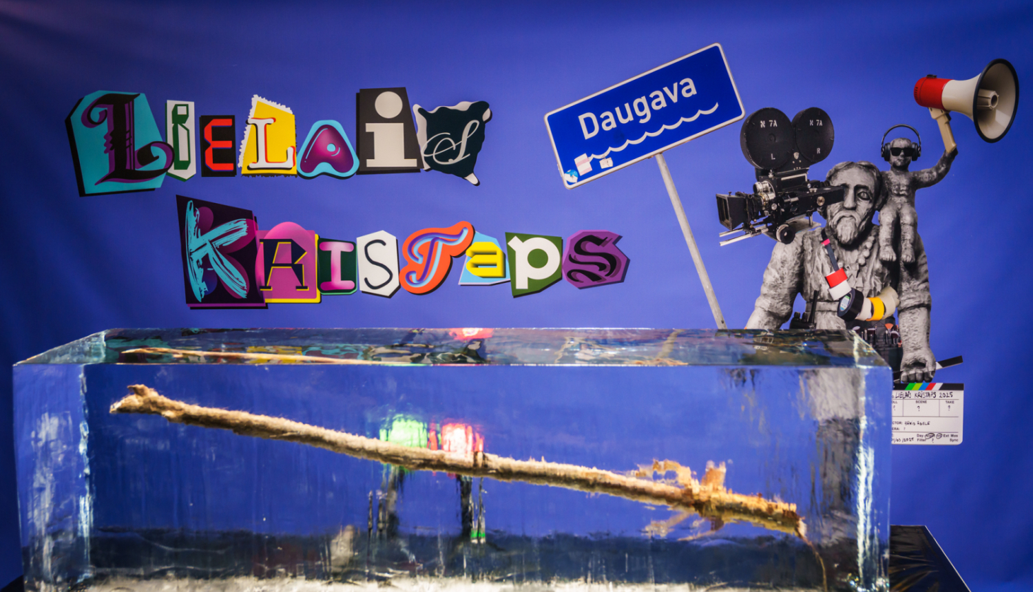 Nacionālās kino balvas „Lielais Kristaps 2025” vizuālā identitāte - baneris ar Lielā Kristapa statueti un ledus gabalā iesalusi koka nūja