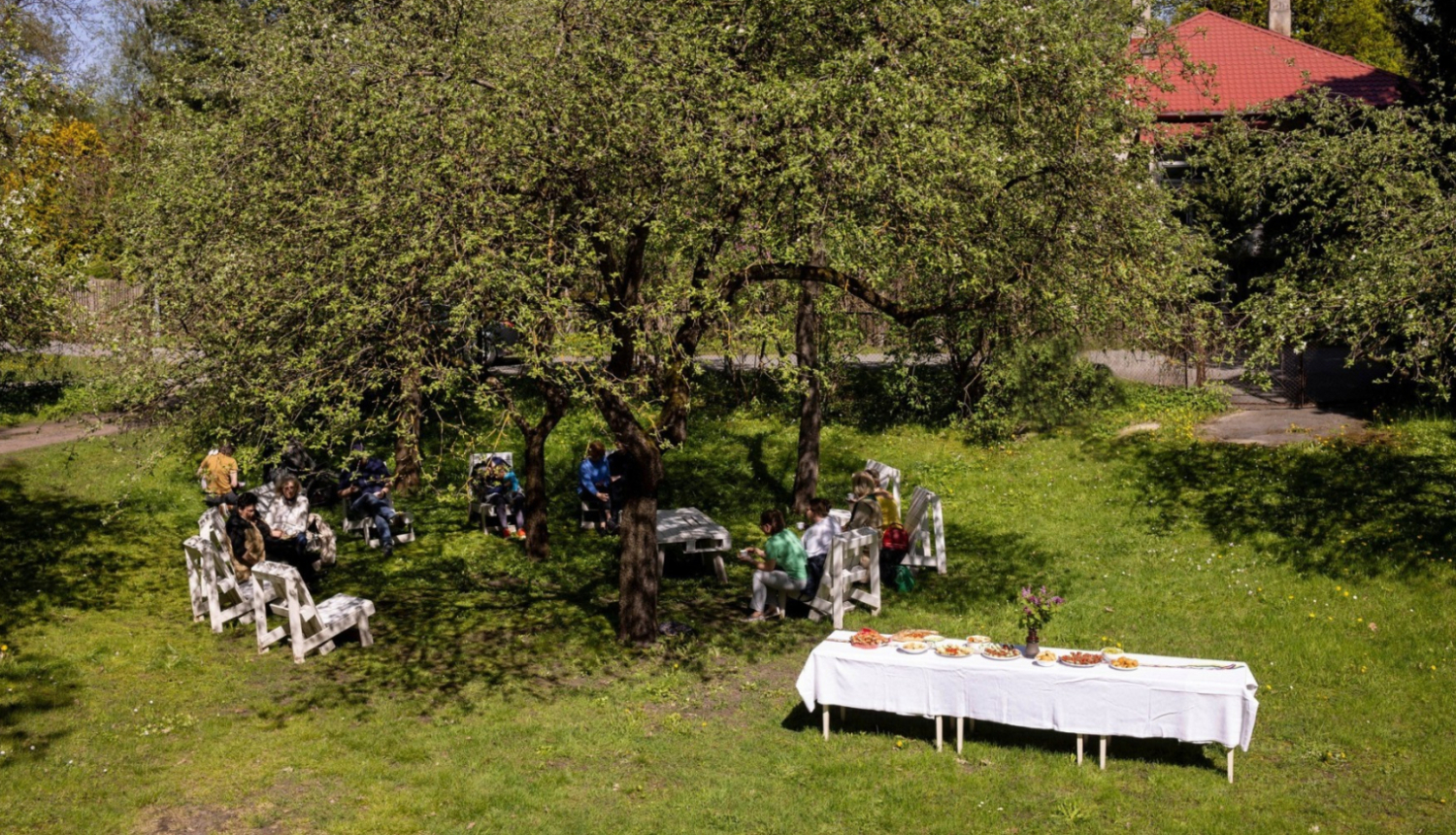 Cilvēki pulcējušies pie balti klāta galda Ojāra Vācieša muzeja dārzā