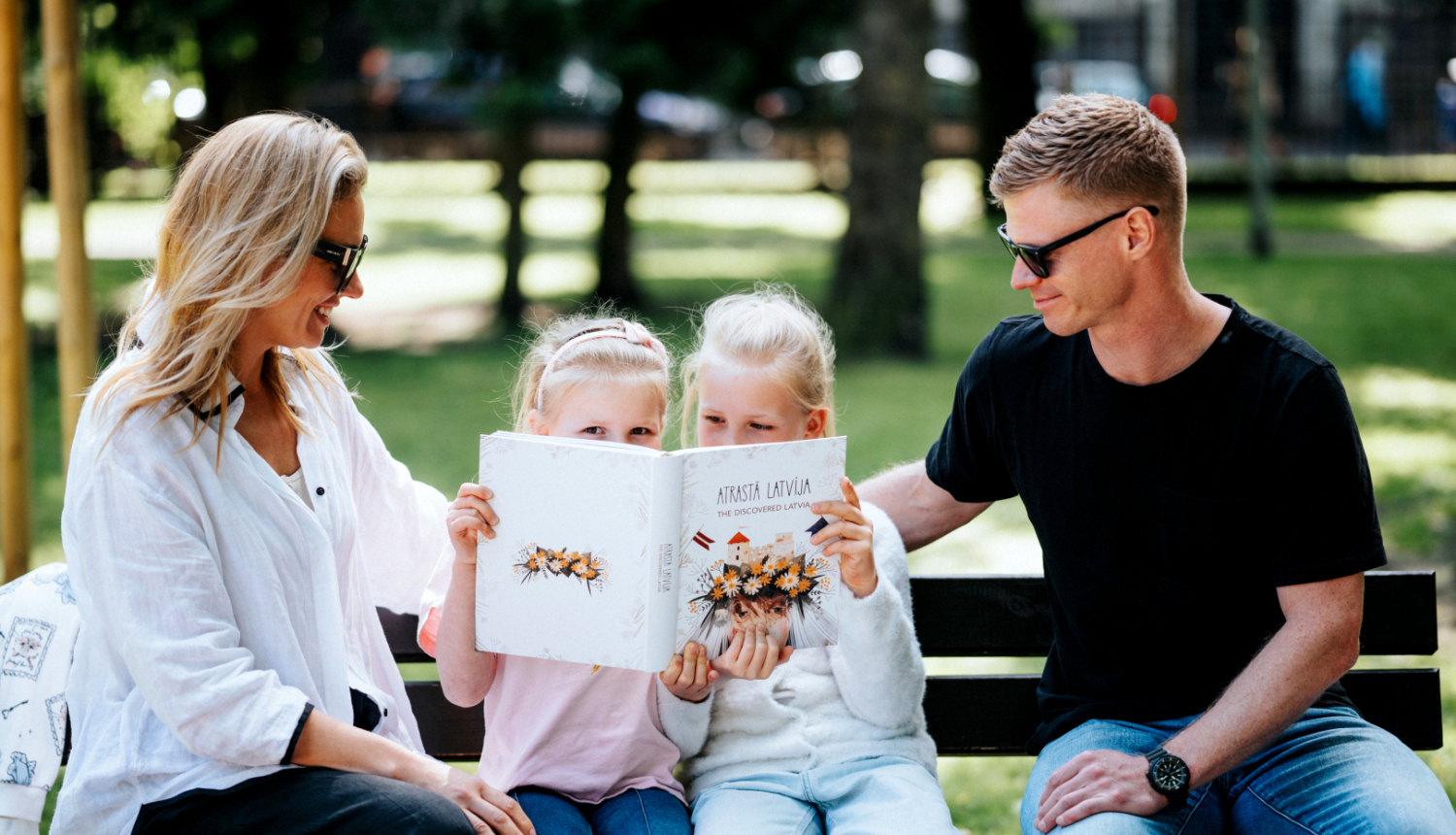 Ģimene - tētis, mamma un divas meitas - brīvā dabā parkā uz soliņa saulainā laikā lasa grāmatu "Atrastā Latvija".