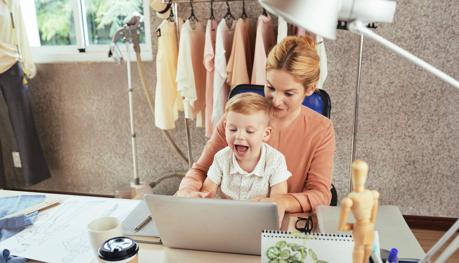 Māte ar bērnu klēpī sēž pie rakstāmgalda un strādā ar portatīvo datora