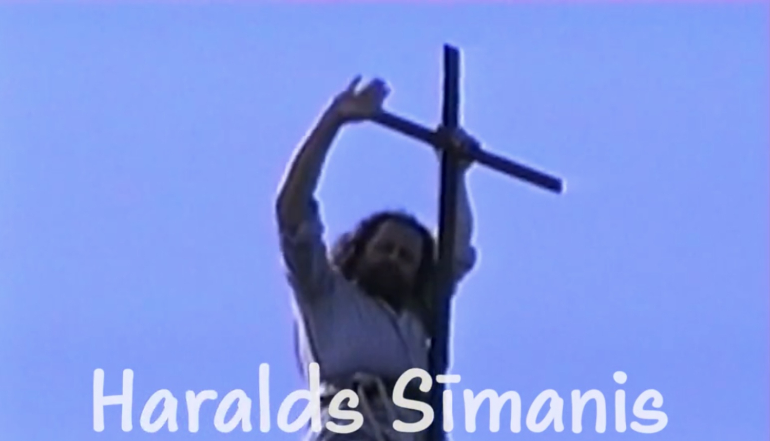 Filmas kadrs ar mūziķi Haraldu Sīmani un koka krustu. Teksts: Haralds Sīmanis, dziesminieks un jumiķis