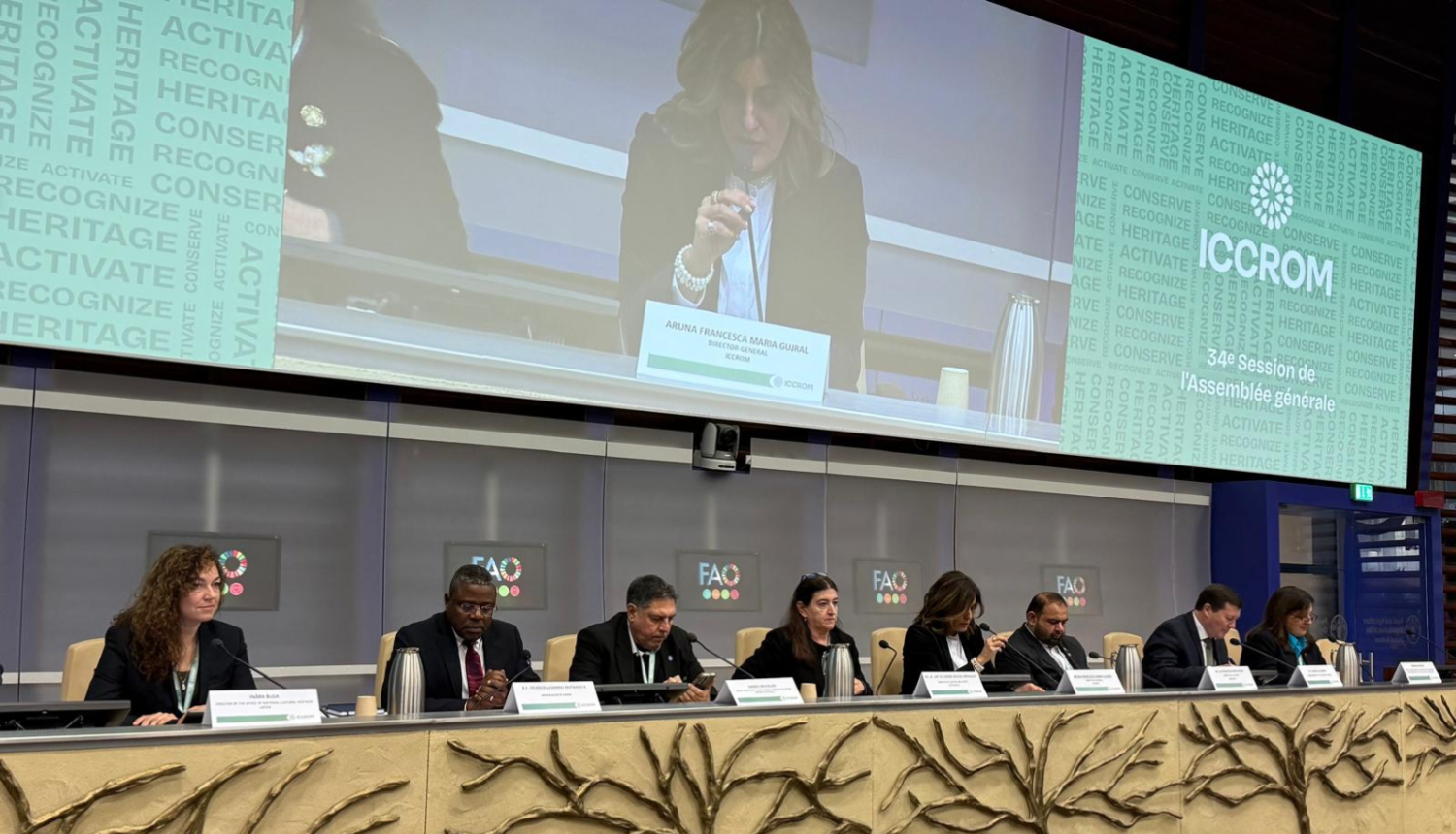 Astoņi cilvēki pie sanāksmju galda, fonā panelis, kur tuvplānā redzams runātājs