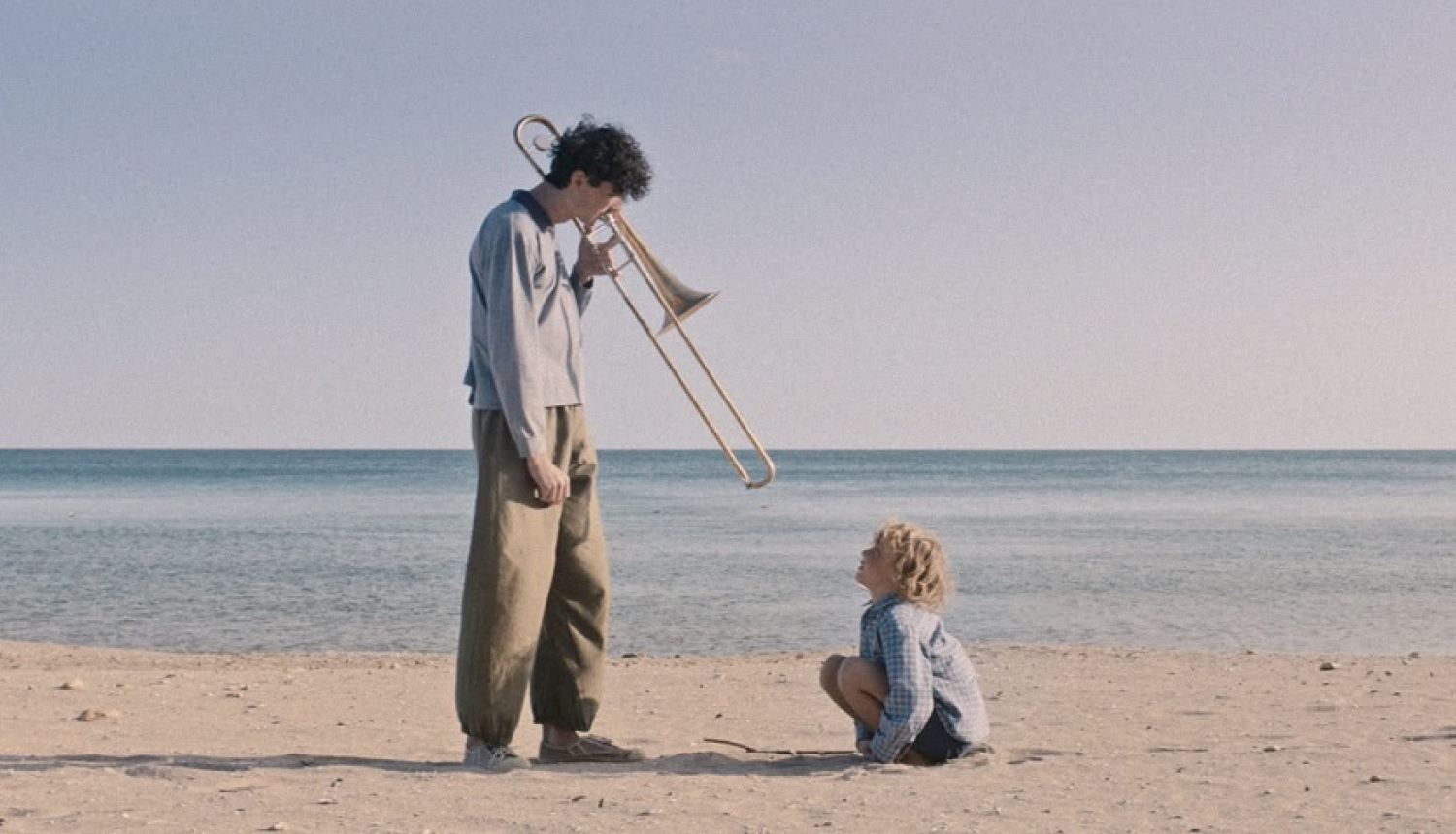 Kadrs no filmas: jūras krastā divi cilvēki - piaugušais ar trombonu rokās un bērns, kas spēlējas smiltīs