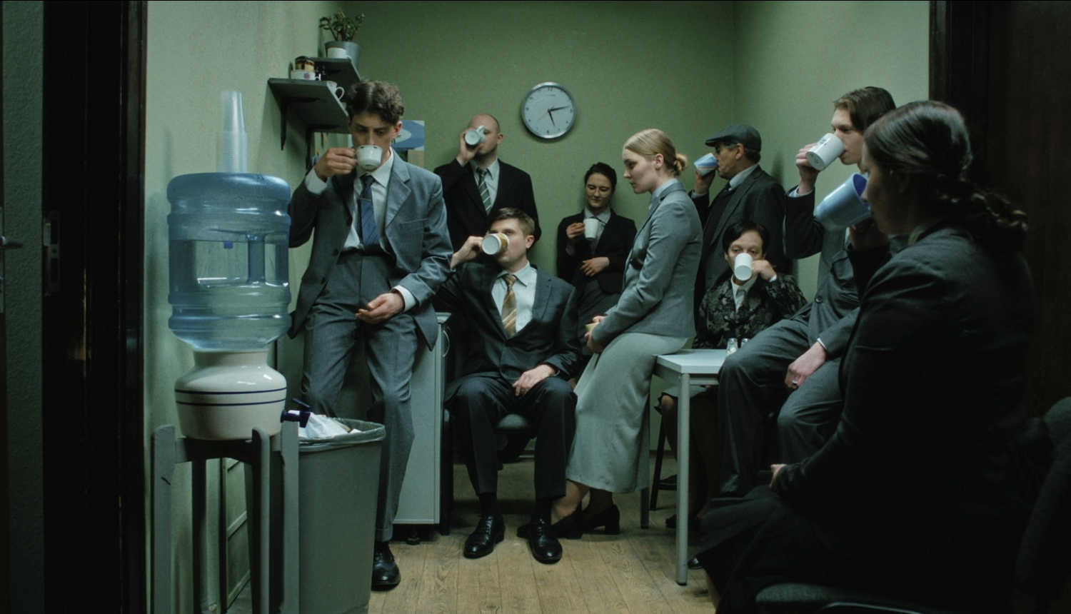 Kadrs no filmas - cilvēku grupa uzvalkos ofisa virtuves telpā