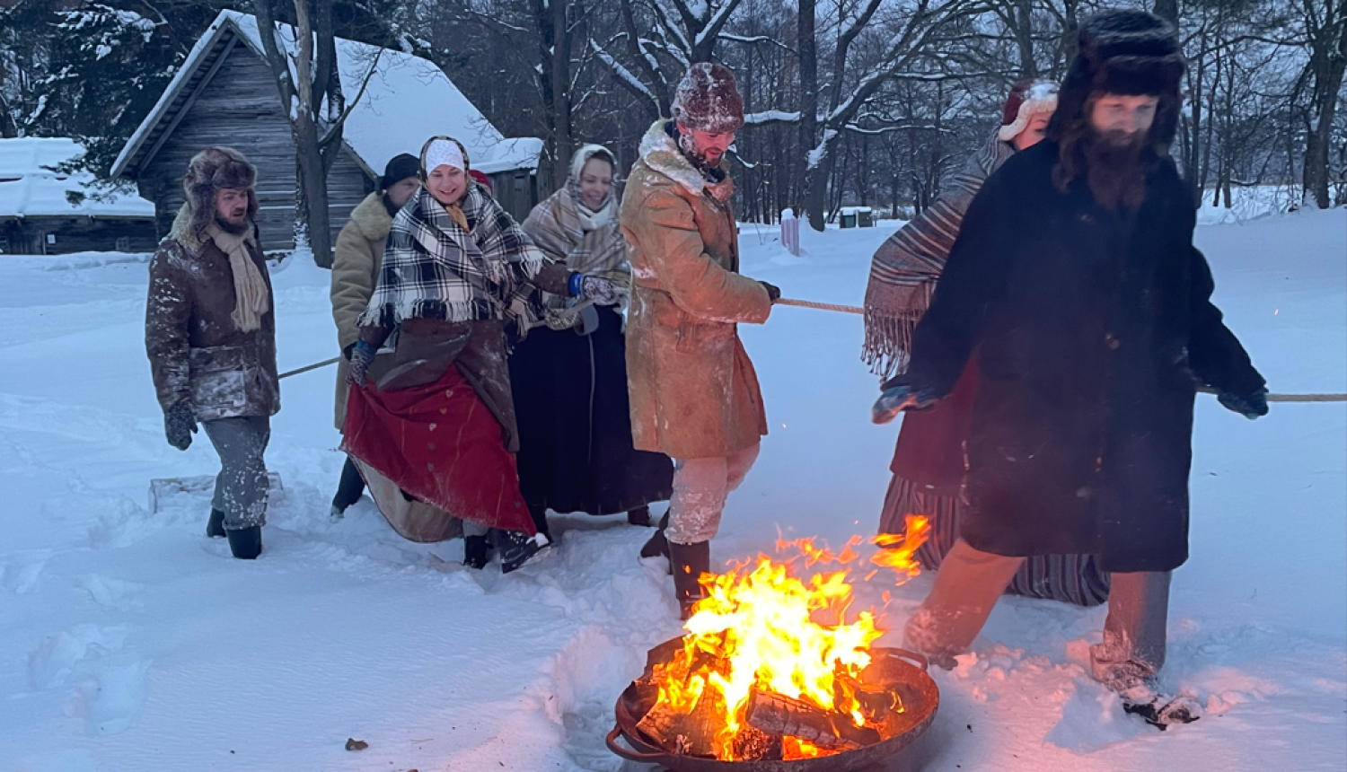 Cilvēku bariņš velk bluķi sniegotā ziemas dienā Brīvdabas muzejā pie ugunskura