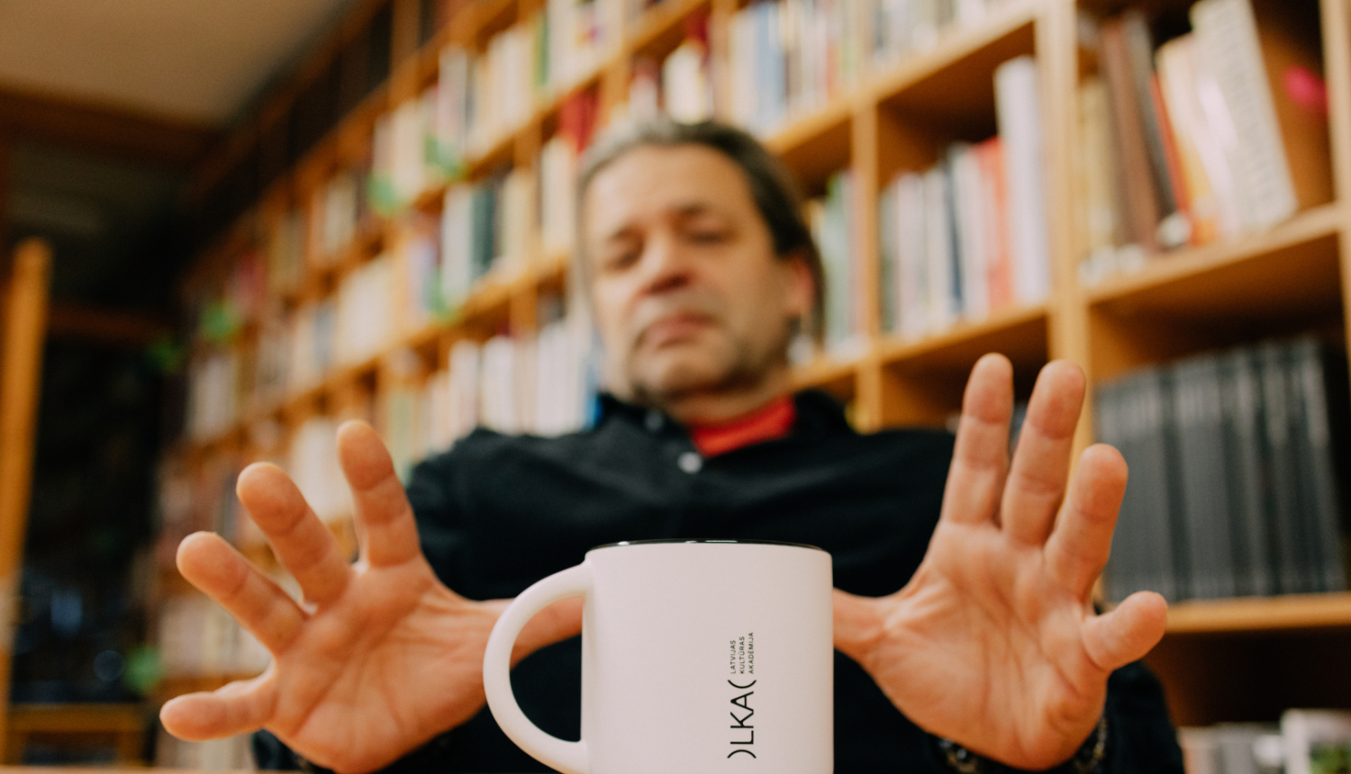Profesors Raimonds Briedis, krūzīte ar Latvijas Kultūras akadēmijas zīmola uzrakstu, fonā grāmatplaukts