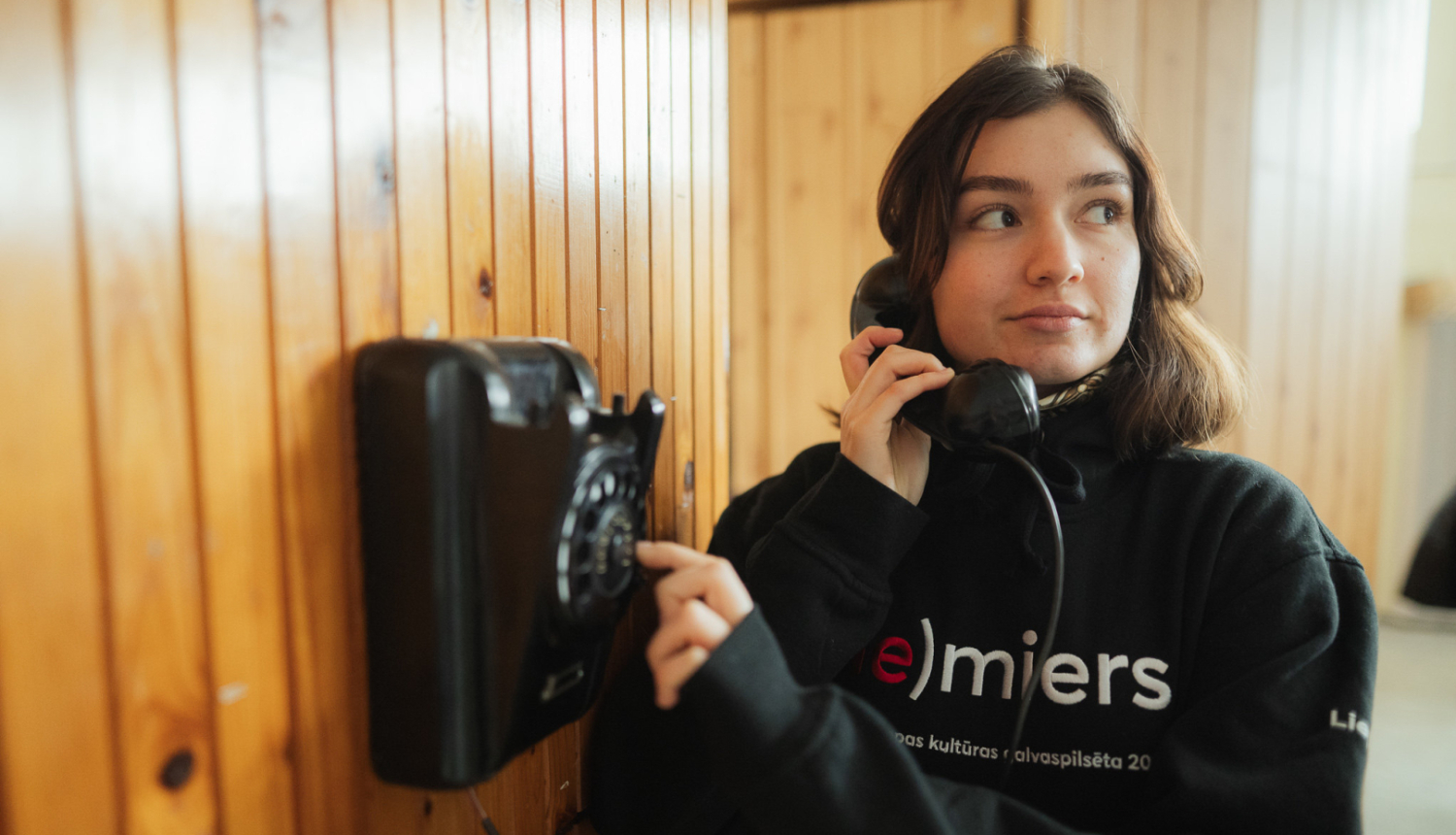 Ilustratīvs attēls ar meiteni, kura stāv pie senlaicīga telefona ar telefona klausuli pie auss un griež ciparnīcu