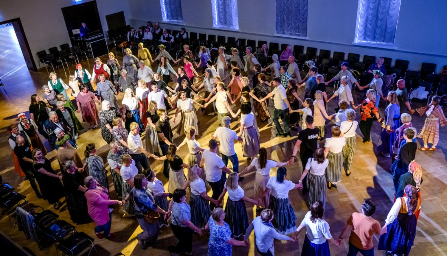 Cilvēki lielā zālē sadevušies rokās, dejo