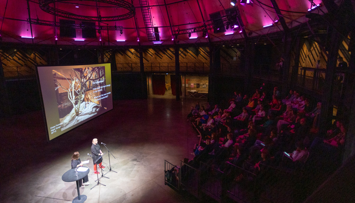 Konferences norise Rīgas cirka arēnā. Divi dalībnieki uzstājas, fonā ekrāns, tribīnēs sēž publika
