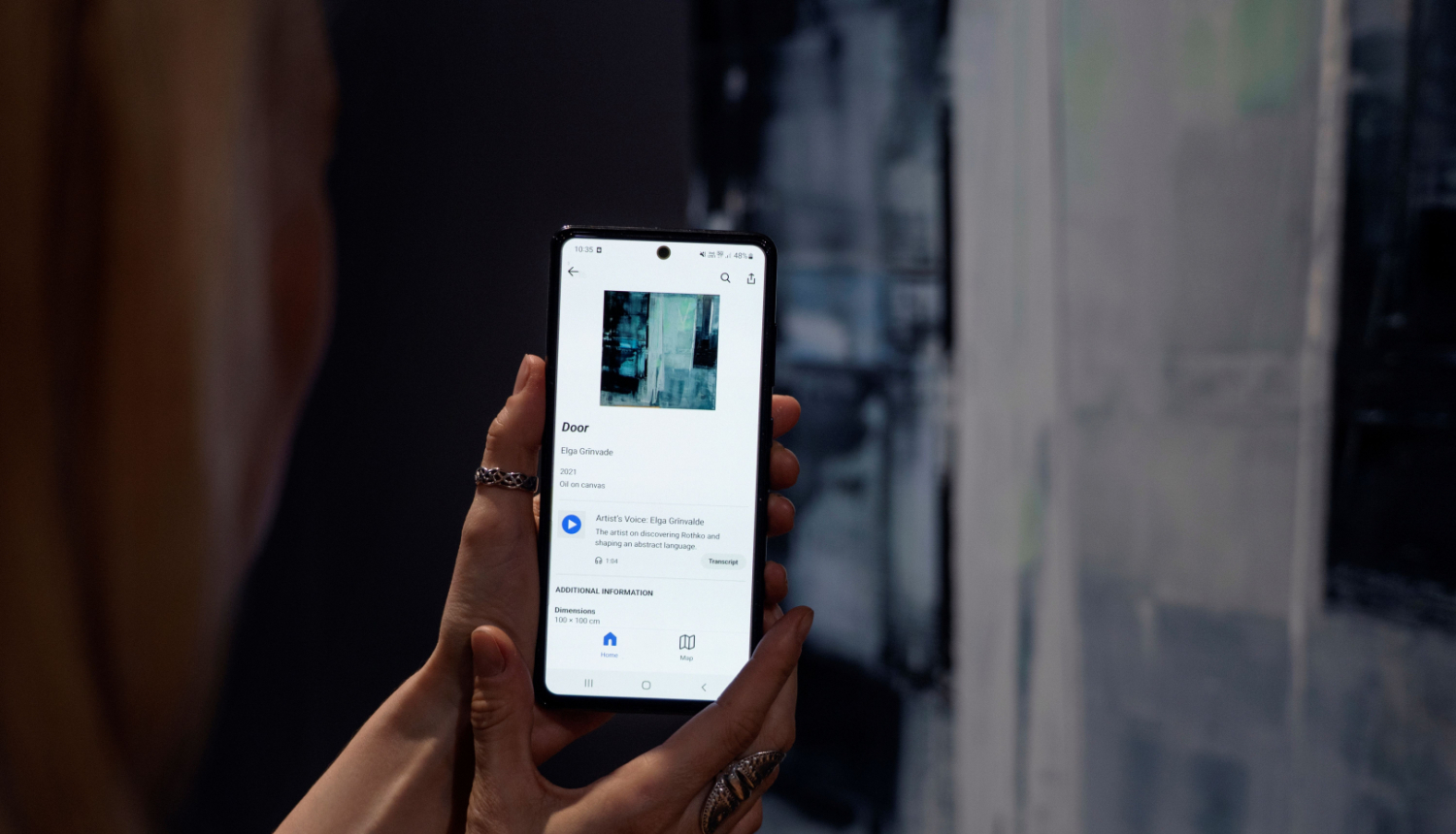 Rokas ar tālruni, kurā atvērta Rotko muzeja mobilā lietotne