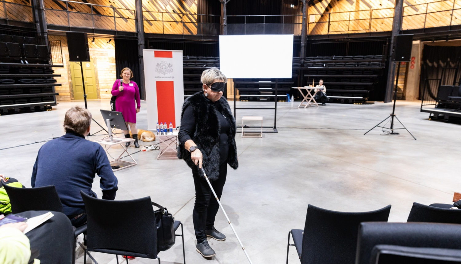 Cilvēks ar spieķi auditorijas priekšā demonstrē piekļūstamības izaicinājumus