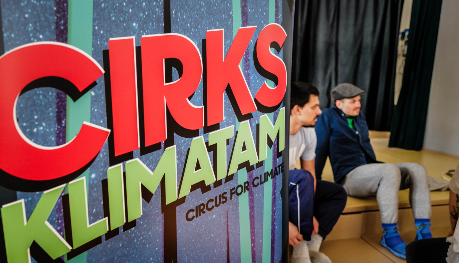 Uzraksts: Cirks klimatam. Divi sēdoši skatītāji.