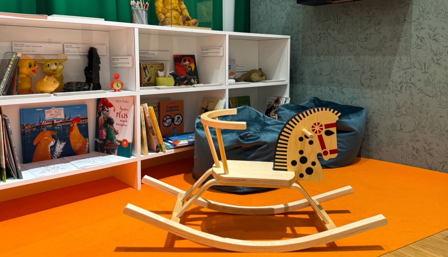 Izstādes iekārtojums plaukti ar bērnu grāmatām, rotaļlietām un koka šupuļzirdziņš
