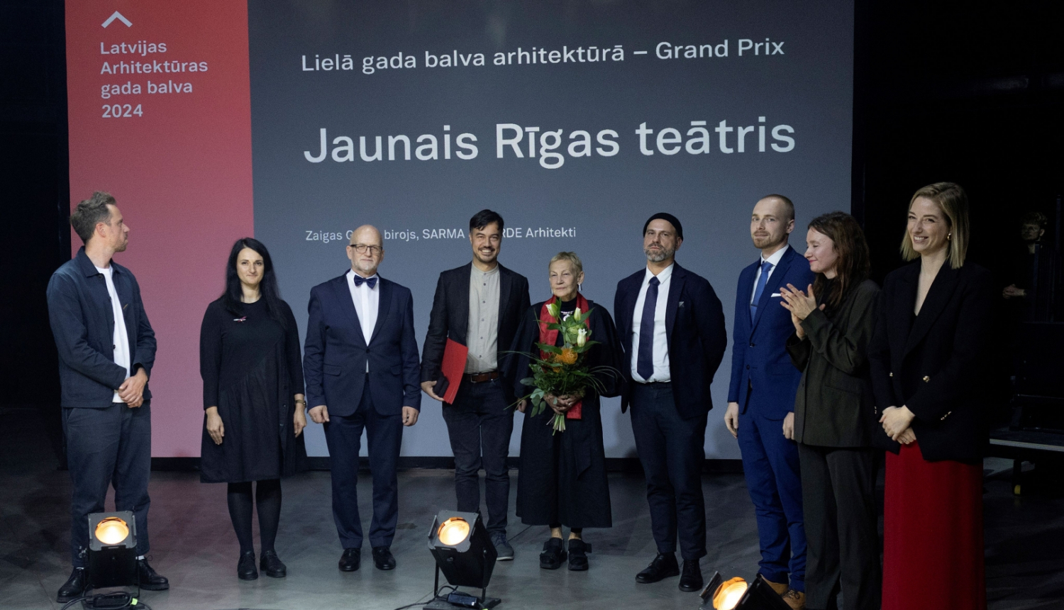 Deviņi cilvēki uz skatuves balvas pasniegšanas laikā