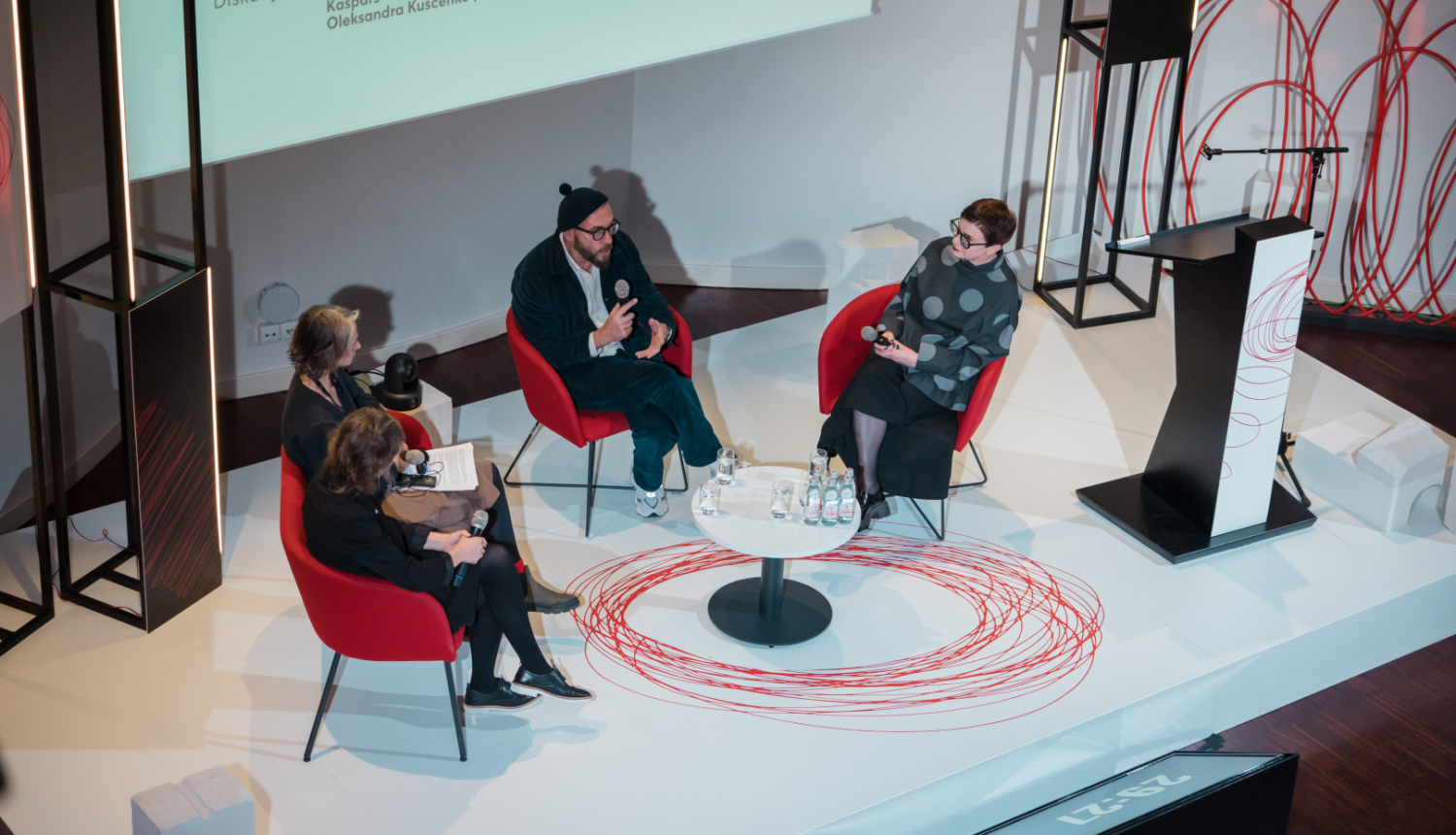 Četri cilvēki sēž modernā konferenču telpā apaļajā diskusijā