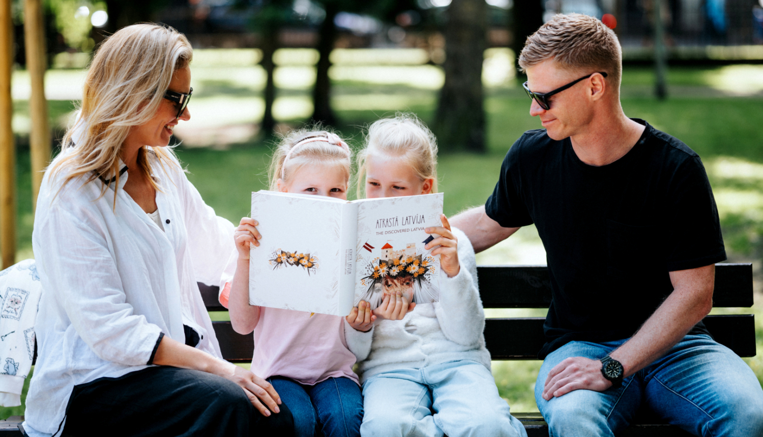 Ģimene - tētis, mamma un divas meitas - brīvā dabā parkā uz soliņa saulainā laikā lasa grāmatu "Atrastā Latvija".