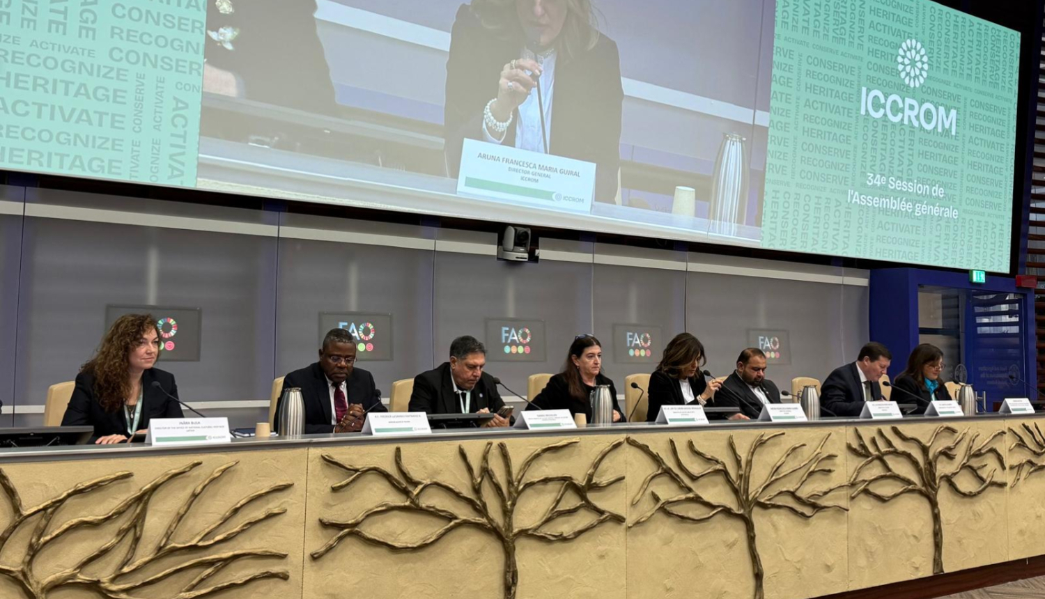 Astoņi cilvēki pie sanāksmju galda, fonā panelis, kur tuvplānā redzams runātājs